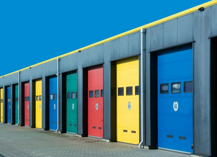 A row of light-industrial storage units with colorful roll-up doors under a blue sky.